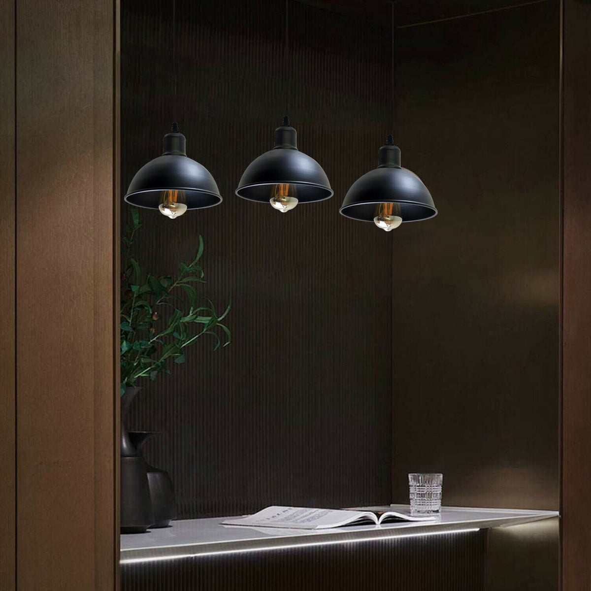Three black pendant lights hanging above a desk with an open book and a glass on a dark wall.