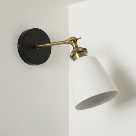 Wall-mounted light fixture with white shade and brass arm on a plain wall.