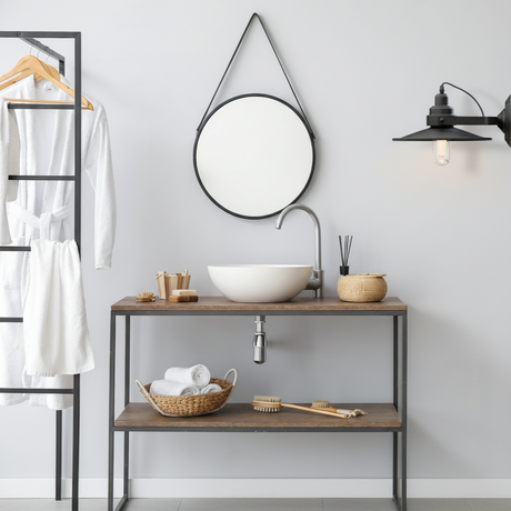 Bathroom interior with a wooden vanity, round mirror, and hanging clothes.